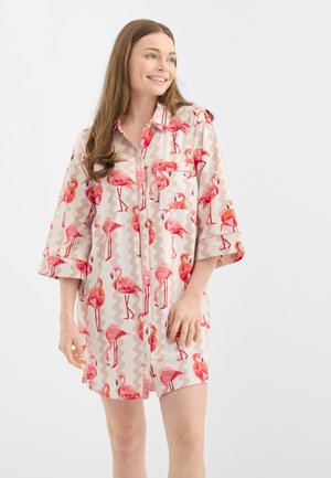 Młoda kobieta uśmiechająca się, ubrana w luźną sukienkę na guziki z motywem różowych flamingów i rękawami do łokcia, na jednolitym tle.