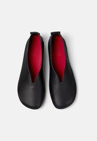 Scarpe slip-on in pelle nera con una texture liscia, dotate di un design a V profondo e soletti rossi in contrasto. Nessuna minuteria o accenti visibili.