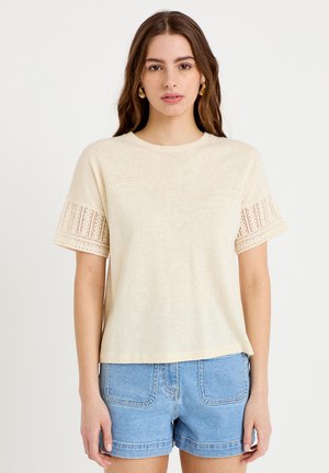 Jeune femme aux longs cheveux bruns portant un haut crème à manches courtes avec des détails en dentelle sur les manches et un short en jean bleu clair, debout dans une posture neutre.