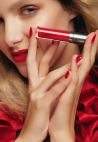 Rød flytende leppestift i en klar tube med sølvhett, holdt mellom hender med lange, røde negler. Dype røde lepper og mykt, bølgete hår er synlig.