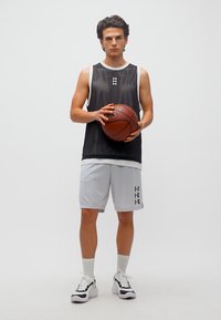 Νεαρός άνδρας με μαύρο αμάνικο αθλητικό φανελάκι και λευκά σορτς μπάσκετ που κρατάει μια μπάλα μπάσκετ, στέκεται μπροστά σε απλό λευκό φόντο.