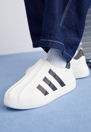Chaussures blanches à enfiler avec un logo trois bandes noir, portées avec des chaussettes grises et un jean bleu foncé, posées sur un sol recouvert d'une moquette bleu clair.