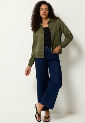 Femme aux cheveux bouclés portant une veste boutonnée vert olive, un top noir, un jean large bleu foncé et des sandales noires, debout sur un fond blanc.