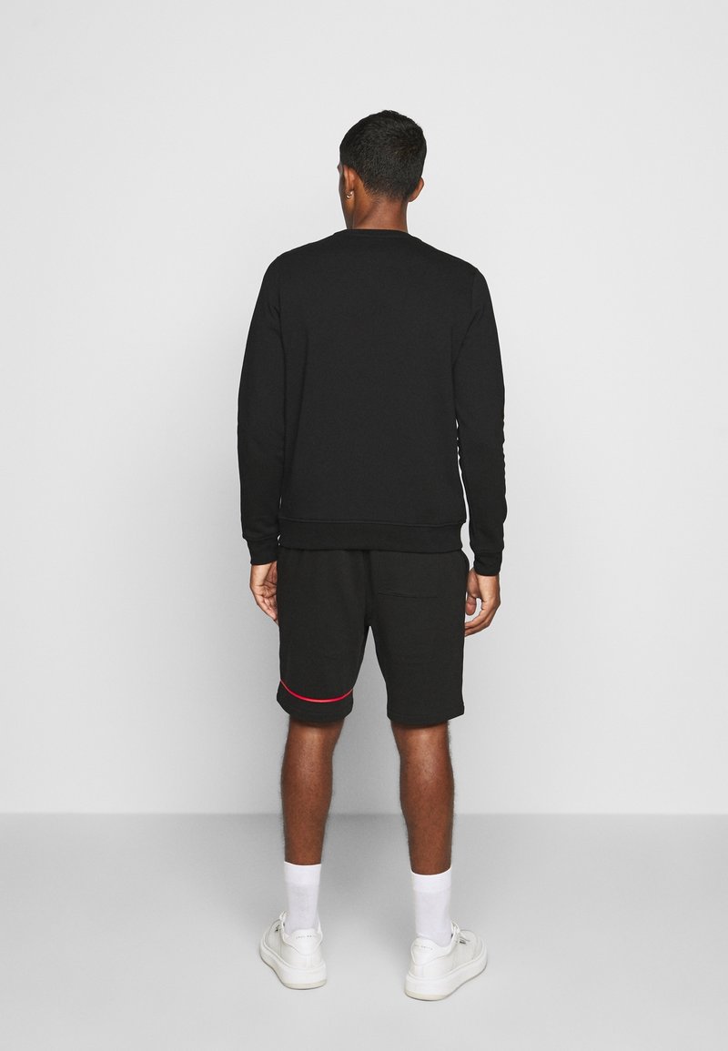Felpa nera con maniche lunghe e polsini a costine, abbinata a pantaloni corti neri con un accento rosso. Sneakers con suole bianche e calze.