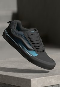 Zwarte en teal sneaker van een glad materiaal, met een ronde neus, een gestructureerde rubberen zool en een opvallend vetersysteem.