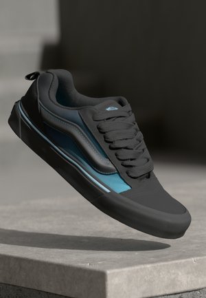 Zwarte en teal sneaker van een glad materiaal, met een ronde neus, een gestructureerde rubberen zool en een opvallend vetersysteem.