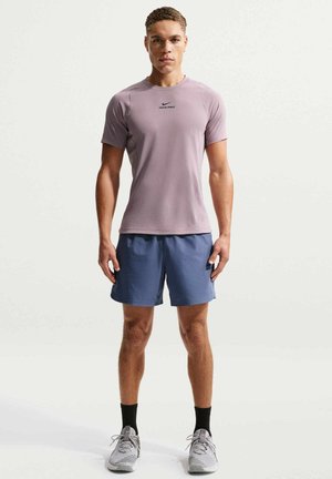 Atletyczny mężczyzna stojący przodem, ubrany w bluzkę Nike Pro w kolorze mauve, niebieskie spodenki, czarne skarpetki i szare buty do biegania na jednolitym tle.