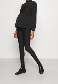 Bluzka ciążowa w czarnym kolorze z teksturowanym koronkowym wykończeniem, w połączeniu z czarnymi błyszczącymi legginsami i czarnymi butami. Prosty, dopasowany krój.