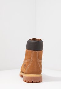 Bottine en daim marron avec un col noir rembourré, des détails de couture blanche et une semelle en caoutchouc texturée, avec un logo sur le côté.