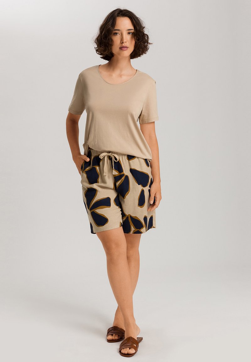 Beige Kurzarmshirt kombiniert mit hellbeigen Shorts, die ein dunkelblaues Blumenmuster und einen Kordelzugbund haben. Braune flache Sandalen sind enthalten.