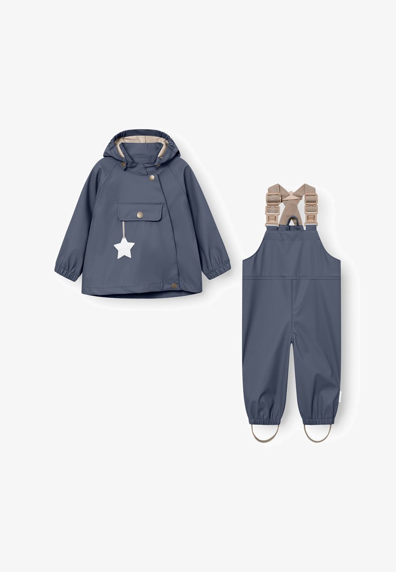 Vaikų tamsiai mėlyna lietaus striukė su gaubtu ir priekinė kišenė, derinama su atitinkamomis neperšlampančiomis užpakalinėmis kelnėmis su reguliuojamomis smėlio spalvos petnešomis ir kojų kilpomis.