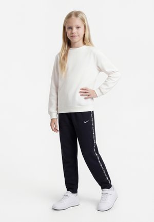Mladé dievča stojí s rukou na boku, má na sebe bielu mikinu, čierne Nike tepláky s logom na pruhu a biele tenisky na bielom pozadí.