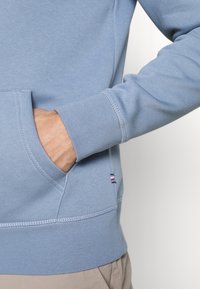 Sudadera azul claro con un bolsillo canguro y puños acanalados. Presenta detalles de costura sutil y un pequeño acento de logo cerca del dobladillo.