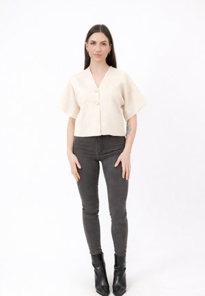 Jeune femme debout portant un haut crème à manches courtes boutonné, un jean slim gris foncé, des bottines noires, avec un tatouage sur l'avant-bras gauche.