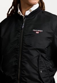 Svart bomberjacka med framsidig dragkedja, ribbad krage och "Polo Sport"-logotyp i röd och vitt. Slät, glansig yta med sidofickor.
