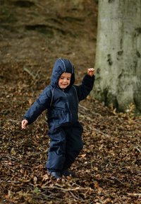 Marinblå, vattentät overall med huva, elastiska muddar och kontrastsömmar, som bärs av ett barn som leker på ett lövtäckt underlag.