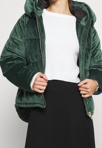 Veste matelassée verte en velours doux, dotée d'une fermeture éclair, d'une grande capuche et de poignets côtelés, portée avec un pull en tricot blanc.