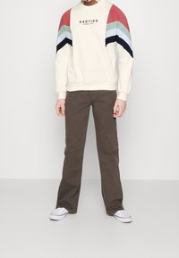 Felpa beige chiaro con dettagli a strisce multicolori in rosso, verde, blu e nero, abbinata a pantaloni a zampa d'elefante marroni e sneaker bianche.