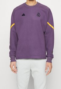 Sudadera morada de manga larga, con cuello redondo, logo negro de adidas, escudo del Real Madrid y una franja amarilla en el hombro.