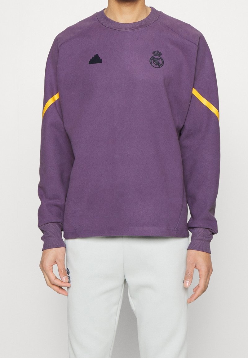 Sudadera morada de manga larga, con cuello redondo, logo negro de adidas, escudo del Real Madrid y una franja amarilla en el hombro.