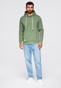 Hombre con sudadera verde, jeans azul claro y zapatillas blancas, de pie contra un fondo claro y liso.