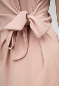 Pale pink dress with a soft, flowing texture, featuring a bow tied at the back, accentuating the waist with draped fabric details.