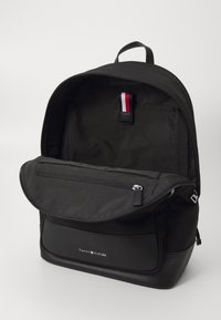 Zaino nero realizzato in tessuto resistente con una finitura liscia. Presenta una tasca frontale con zip e spallacci regolabili. Logo visibile nella parte bassa anteriore.