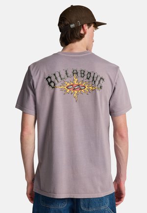 Jonge man draagt een stoffig paarse Billabong t-shirt met een vurige zon grafiek en logo op de achterkant, gecombineerd met een blauwe spijkerbroek en een bruine pet.