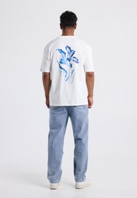 White cotton t-shirt featuring a blue floral design on the back. Model wears light blue denim jeans and white sneakers.