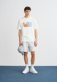 Fehér rövid ujjú póló színes "AIR JORDAN" grafikával, szürke rövidnadrággal, fehér sportcipővel és szürke sapkával párosítva. Egyszínű háttér.