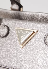 Borsa a tracolla argentata con texture, caratterizzata da un logo triangolare Guess con accenti dorati. Include dettagli neri e una struttura elegante.