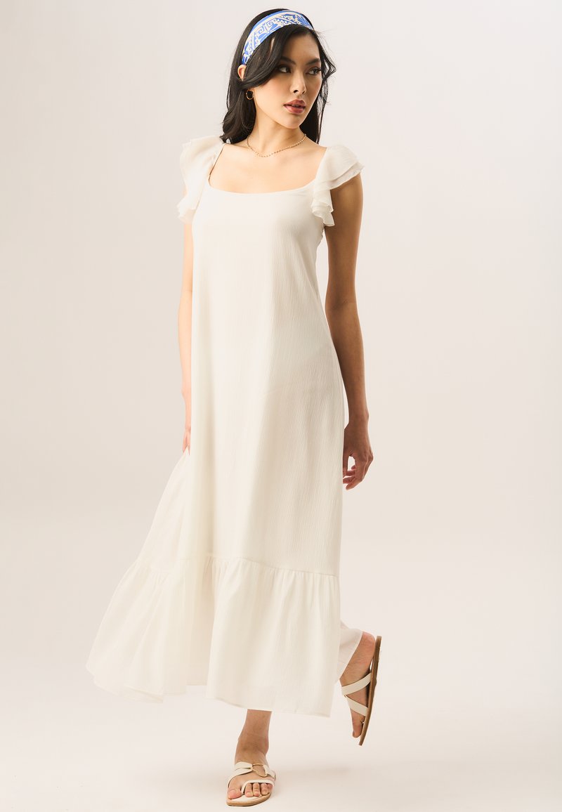 White maxi dress with ruffled sleeves, square neckline, and tiered hem. Worn with light-colored sandals. Smooth texture, lightweight fabric.