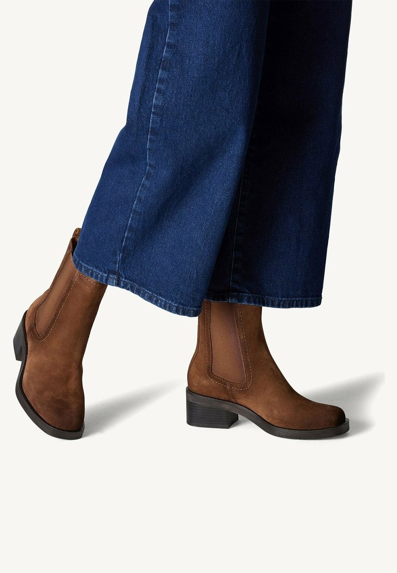 Bottes cheville en daim marron avec panneaux latéraux élastiques, portées avec un jean large bleu foncé, sur un fond blanc uni.