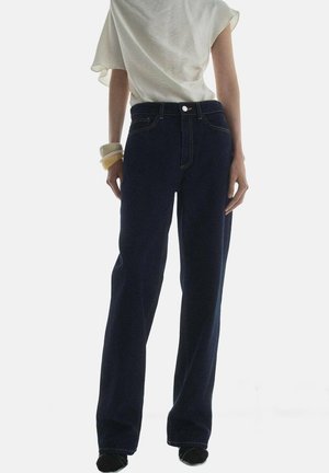 Donkerblauwe denim wijduitlopende jeans met contrasterende gele stiksels, een sluiting met een voorknop en zijzakken. Ggedragen met een lichtgekleurde top.