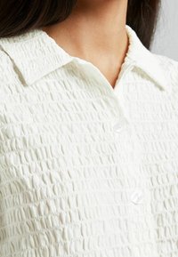 White textured blouse featuring a crinkled design, button closure, and a classic collar. The fabric has a soft, lightweight appearance.