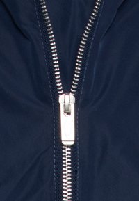 Marinblå jacka med en silverfärgad dragkedja i mitten, med synliga tänder och en märkesdragkedjepull, mot en slät tygtextur.