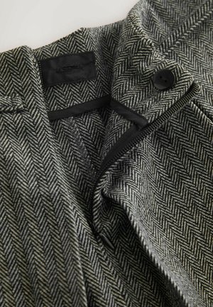 Stoff mit Fischgrätmuster in Schwarz und Weiß, ausgestattet mit Reißverschluss und Knopfverschluss. Glatte Textur mit maßgeschneidertem Design und schwarzen Akzenten.