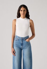 White sleeveless bodysuit with a mock neck, paired with high-waisted light blue wide-leg jeans. Smooth fabric with minimal design details.