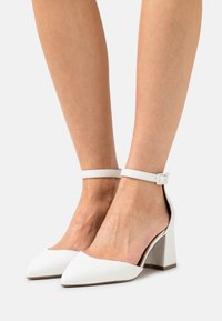 Chaussures à talons hauts blanches avec un bout pointu et une sangle de cheville. Fabriquées en matériau lisse avec un talon bloc solide et un design minimaliste.