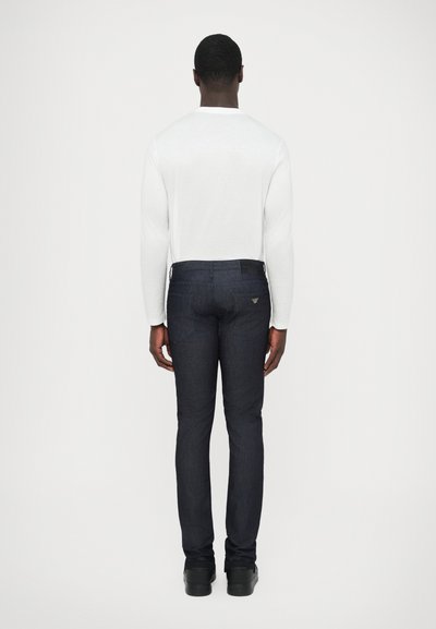 Σκούρο μπλε τζιν παντελόνι με στενή εφαρμογή, με πίσω τσέπες και διακριτικό λογότυπο, συνδυασμένο με εφαρμοστή λευκή μακρυμάνικη μπλούζα.