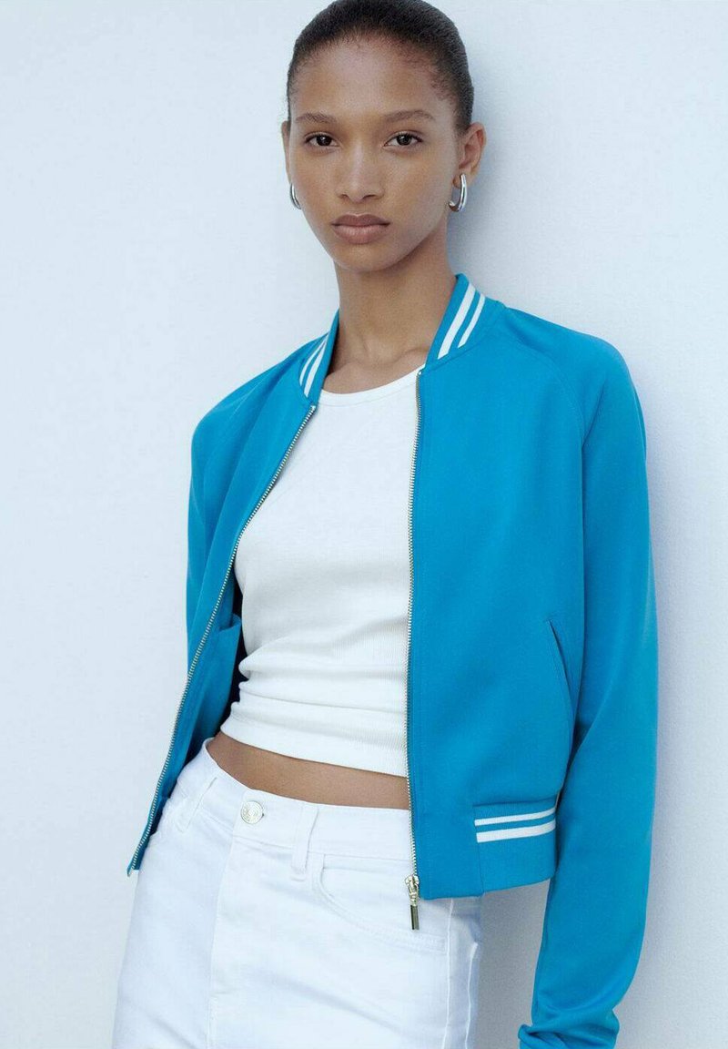 Giubbotto bomber turchese con strisce bianche sul colletto e sui polsini, abbinato a un crop top bianco e jeans bianchi a vita alta.