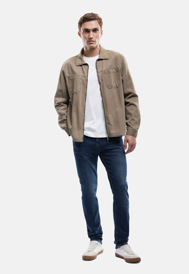 Vaaleanruskea puuvillatakki, jossa on kaksi rintataskua, on puettu valkoisen t-paidan päälle. Kuvassa näkyvät myös siniset farkut ja valkoiset lenkkitossut, joissa on ruskeat pohjat.