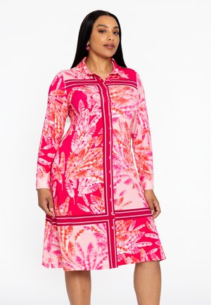 Robe chemise à imprimé feuilles tropicales en rose et corail, avec des manches longues et une jupe légèrement évasée. Texture du tissu lisse.