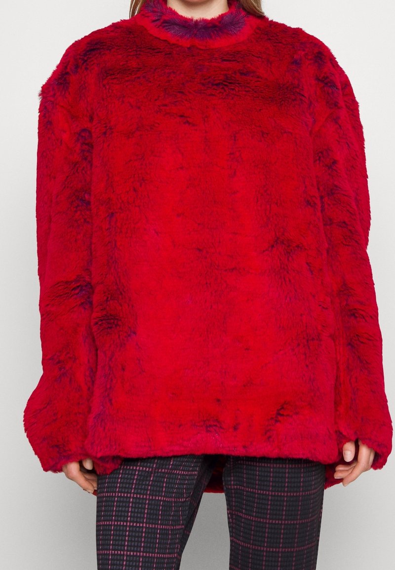 Pull rouge en peluche surdimensionné avec un col montant et une surface texturée. Les manches amples dépassent les poignets. Associé à un pantalon noir à motifs.
