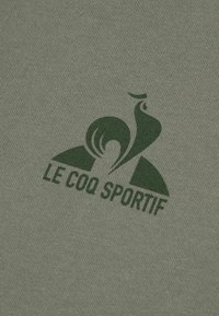 Tkanina olivově zeleného trička s hladkým povrchem, na kterém je tmavě zelené logo kohouta a text "LE COQ SPORTIF" pod ním.