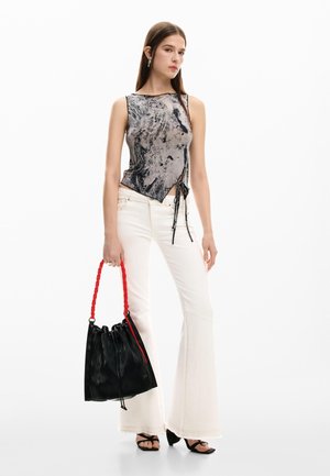 Femeie purtând un top fără mâneci cu imprimeu abstract, blugi evazați albi, sandale negre, ținând o geantă neagră texturată cu mânere roșii.
