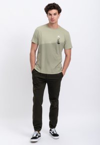 T-shirt verde oliva con stampa grafica in alto a sinistra, abbinata a pantaloni jogger verdi scuri e sneakers nere con suola bianca.