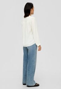 Chemisier blanc à manches longues avec une coupe décontractée, présentant un dos smocké et des poignets volantés. Associé à un jean bleu clair à jambes larges et des sandales noires.