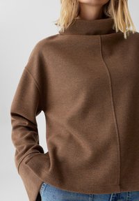 Pullover in maglia marrone con colletto alto, caratterizzato da un dettaglio sulle cuciture laterali e maniche rilassate. Il materiale ha una consistenza morbida e una leggera lucentezza.