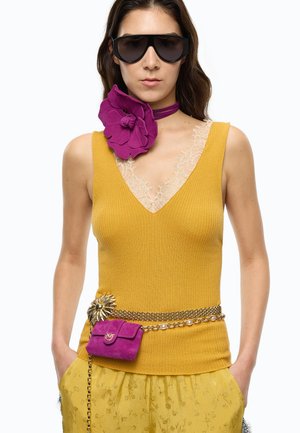 Femme portant de grandes lunettes de soleil noires, un top jaune moutarde sans manches avec un bord en dentelle, un collier ras du cou à fleur violette et une ceinture chaîne dorée avec une pochette violette.